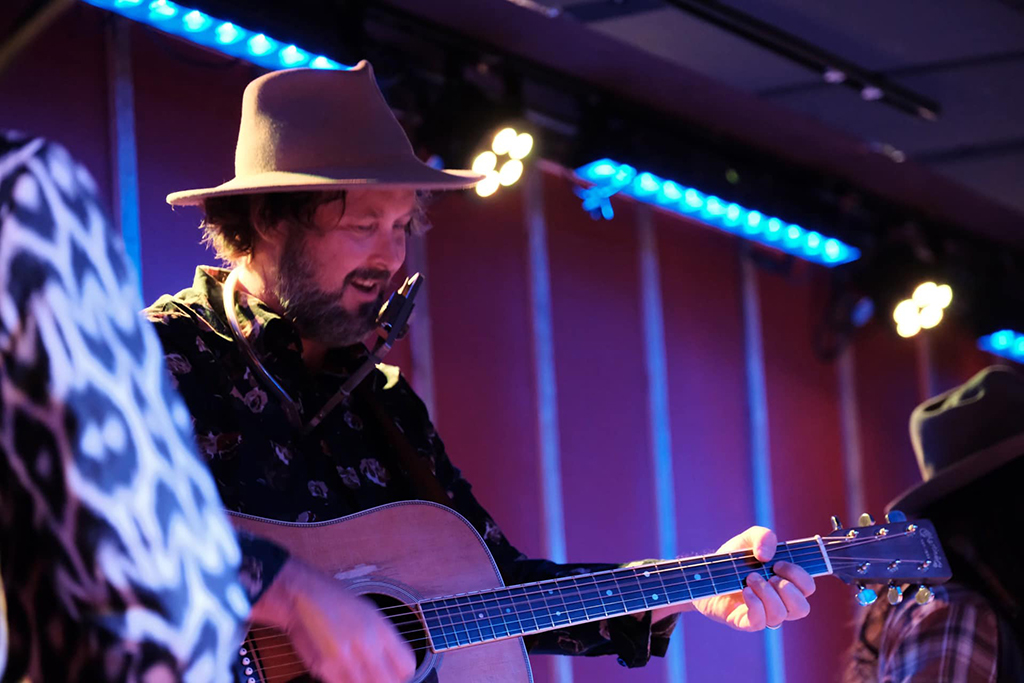 This is a photo of a musician playing an acoustic guitar on a stage. The musician is wearing a hat and a patterned shirt. The background consists of a stage with blue lighting and a red curtain. There is another musician in the background, also wearing a hat.