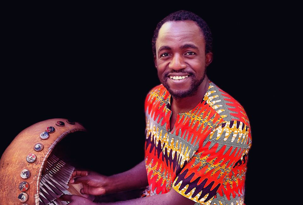 This is a photo of a person playing a traditional African instrument. The person is wearing a colorful shirt with a red, orange, and yellow pattern. The instrument is a wooden harp-like instrument with strings and small metal discs attached to the top. The background is black.