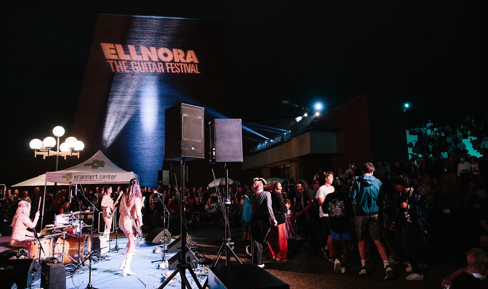 An all woman band plays on a stage in an outdoor ampitheatre. ELLNORA is projected onto the adjacent building.