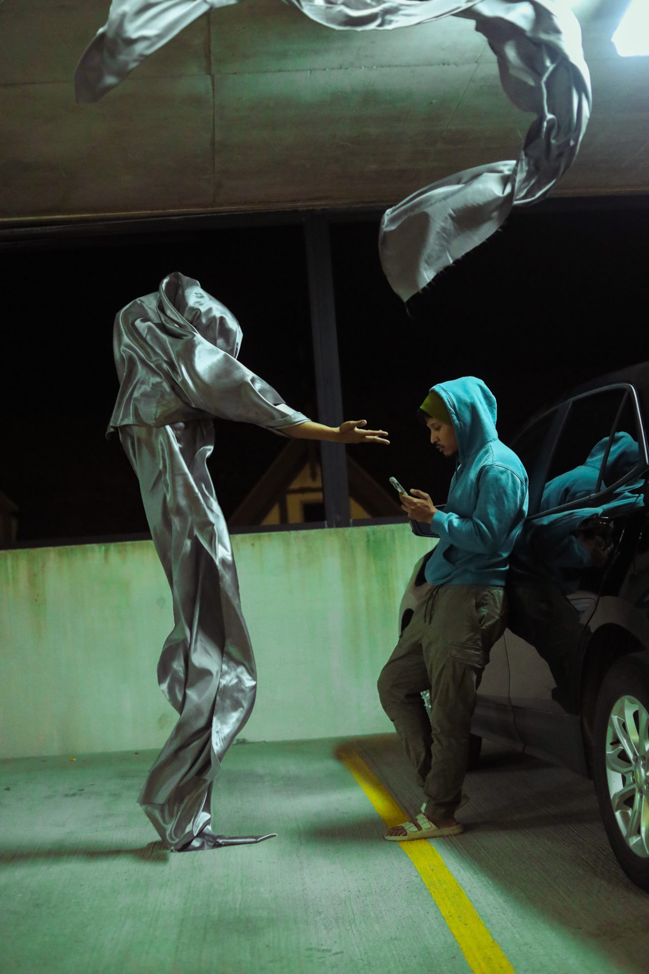 Marcus Flinn stands in a parking garage leaning against a car while on his phone. There is a large figure looming over him made up of silver fabric with an arm extended towards him. 