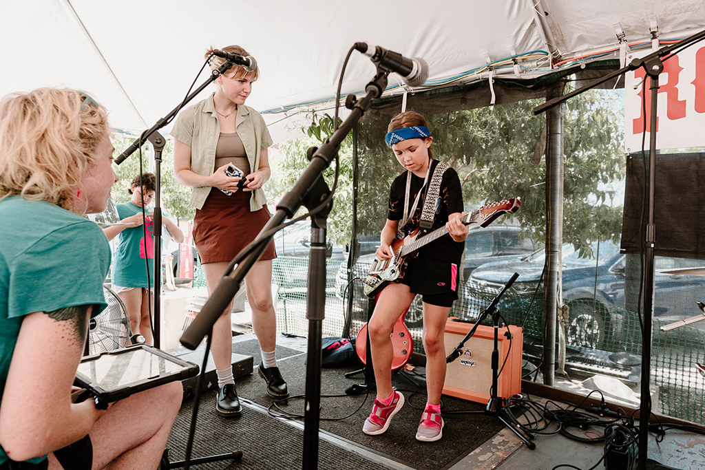 This is a photo of a band performing on a stage under a tent. The stage is set up with microphones, amplifiers, and other musical equipment. The band members are playing a guitar, a bass guitar, and a microphone. The background consists of trees and a parking lot.