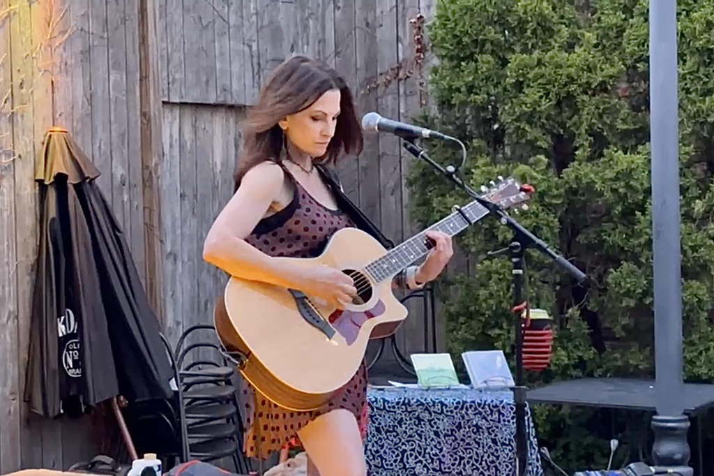 This is a photo of a person playing guitar and singing into a microphone on an outdoor stage. The person is wearing a brown dress with a floral pattern and is playing a light-colored acoustic guitar. The stage is made of wood and there is a black umbrella and a table with a blue tablecloth in the background. The photo was taken during the day and there are trees in the background