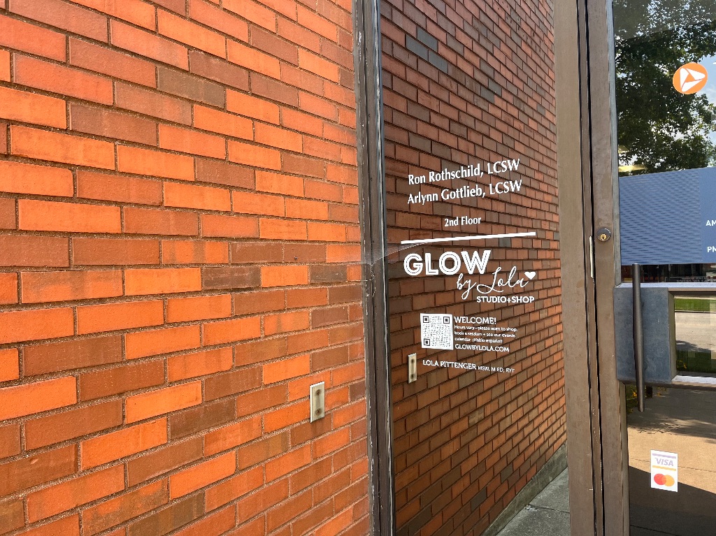 The entry way to Glow by Lola. A large glass door connects to a red brick building. Glow by Lola is printed on the window. 