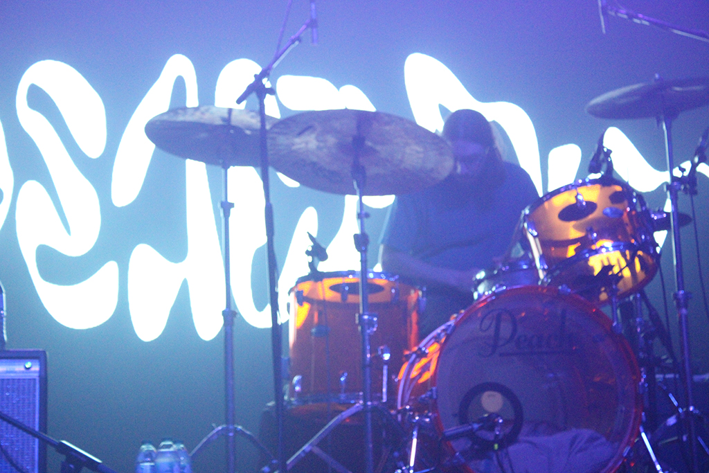 This is a photo of a drummer playing on a stage with a blue light and a white neon sign in the background. The drummer is playing on a drum set with a bass drum, a snare drum, a floor tom, and two cymbals. The bass drum has a logo that reads “Pearl”. The white neon sign in the background is in the shape of a wave and reads “Peach Pit”. The stage is lit with a blue light, creating a moody atmosphere.