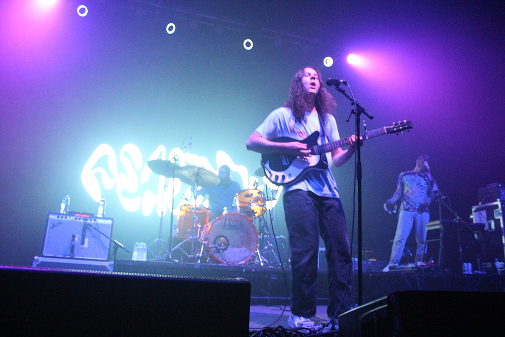 This is a photo of a band performing on a stage with purple lighting. The stage has a large banner with the band’s name written in a cursive font. The band members are playing various instruments, including a guitar, drums, and a keyboard. The faces of the band members are blurred to protect their privacy. The foreground of the photo shows a speaker and part of the stage barrier.