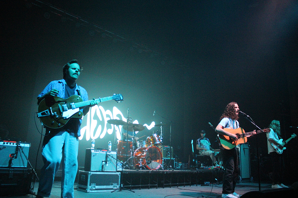 This is a photo of a band performing on a stage in a concert venue. The stage is lit with green and blue lights. The band members are playing various instruments such as guitars, drums, and keyboards. The band’s name, “Hoss”, is written in large white letters on the backdrop.