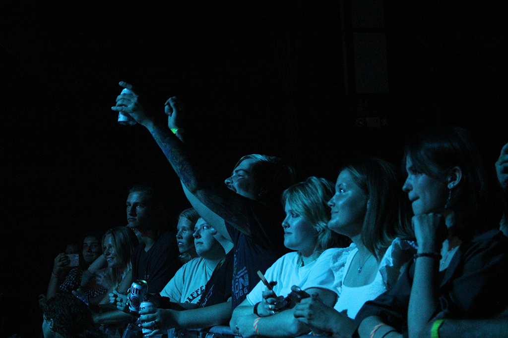 This is a photo of a group of people at a concert or event. The photo is taken from the side and shows the people in the front row. The people are holding up their phones and taking photos or videos. The background is dark and the people are lit up by the stage lights.