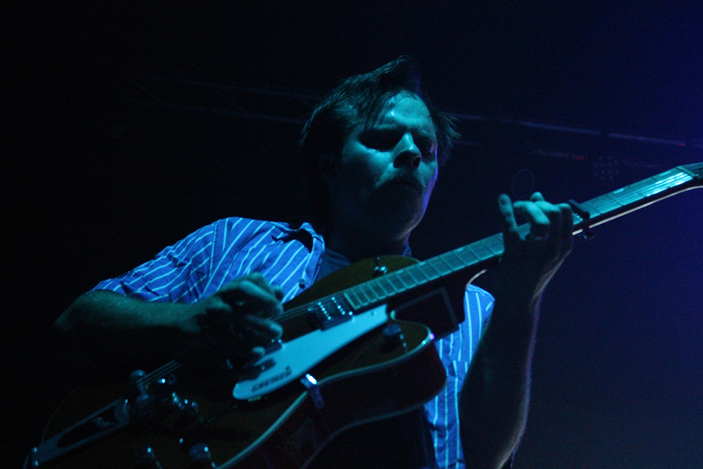 This is a photo of a person playing a guitar on a stage. The person is wearing a striped shirt and is playing a black and white guitar. The background is dark with blue lighting.
