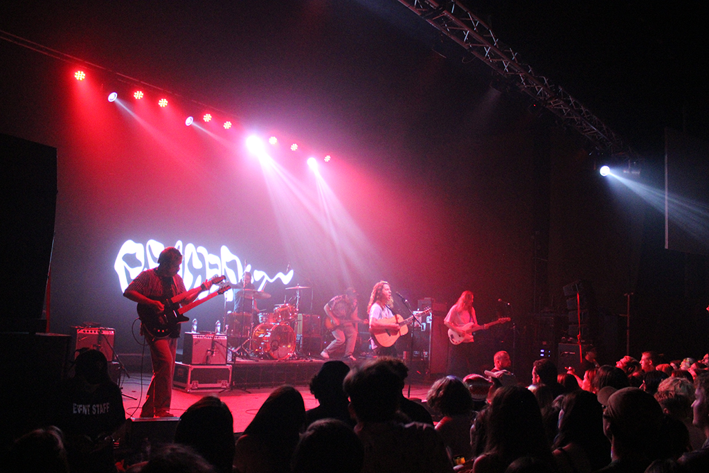 This is a photo of a rock concert in a dark venue with a stage and a crowd. The stage is lit with red and white lights and has a large banner with the band’s name “Peach Pit” in white letters. The band members are playing their instruments on the stage, with the lead singer in the center. The crowd is standing in front of the stage, with some people holding up their phones to take photos or videos. The venue has a high ceiling with metal beams and a large speaker system.