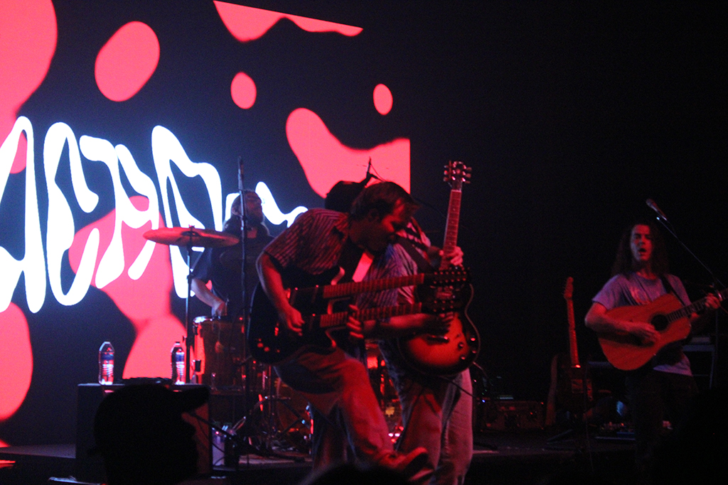 This is a photo of a band performing on a stage with a red and black background. The background is a large screen with red and black abstract shapes and lines. The band members are playing guitars and a drum set. They are wearing casual clothes. The stage is lit with spotlights, and the photo is taken from the audience’s perspective.