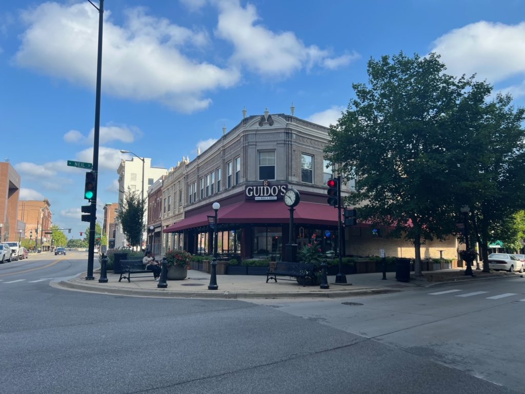 Guido's Bar & Grill in Downtown Champaign has a maroon awning and outdoor seating behind black planters. The exterior of Guido's is pictured on a summer's day with blue skies and fluffy clouds, and the trees have green leaves. Photo by Alyssa Buckley.