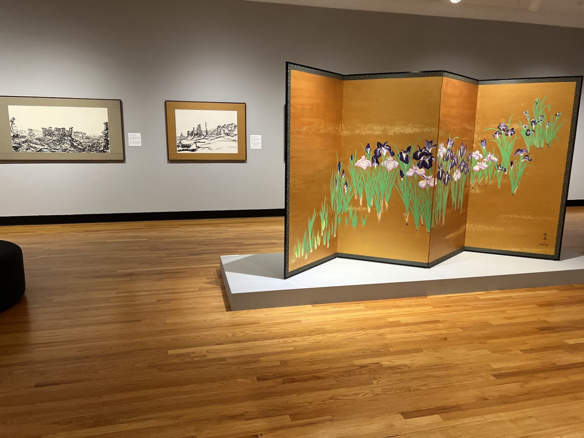 A gallery-wide shot of the Shozo Sato exhibit; a large screen is in the center of the room with a cognac background and flowers; black and white art work hangs on the back wall.