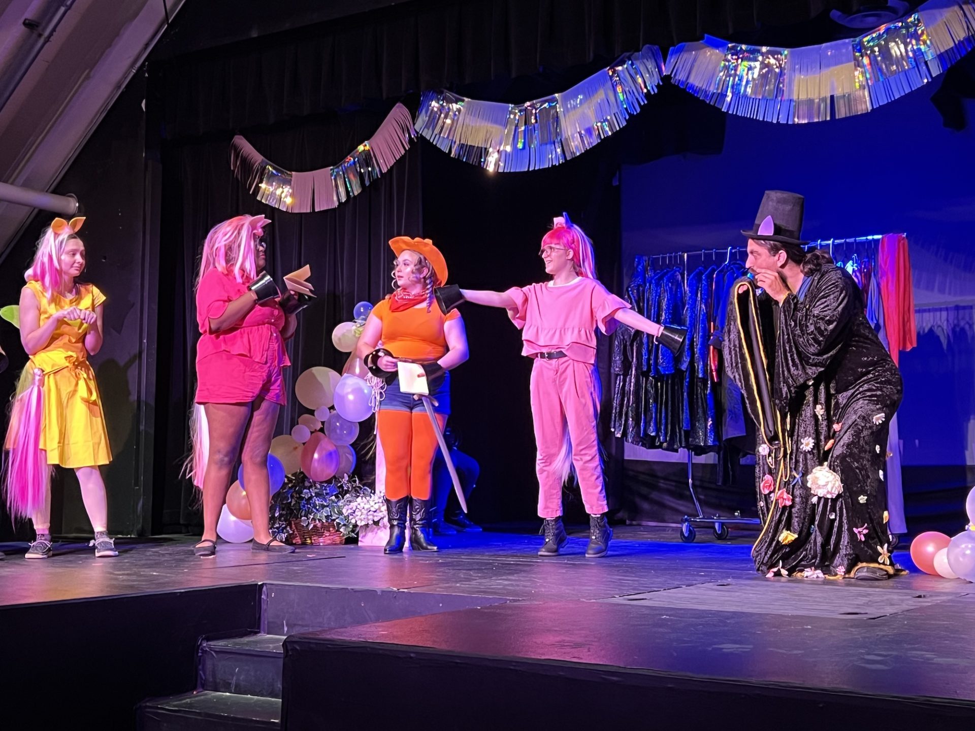 A darkly lit theatre stage; there are party decorations; actors stand on stage dressed as colorful magical ponies. A man is on the right, with a black hat and dressed all in black.