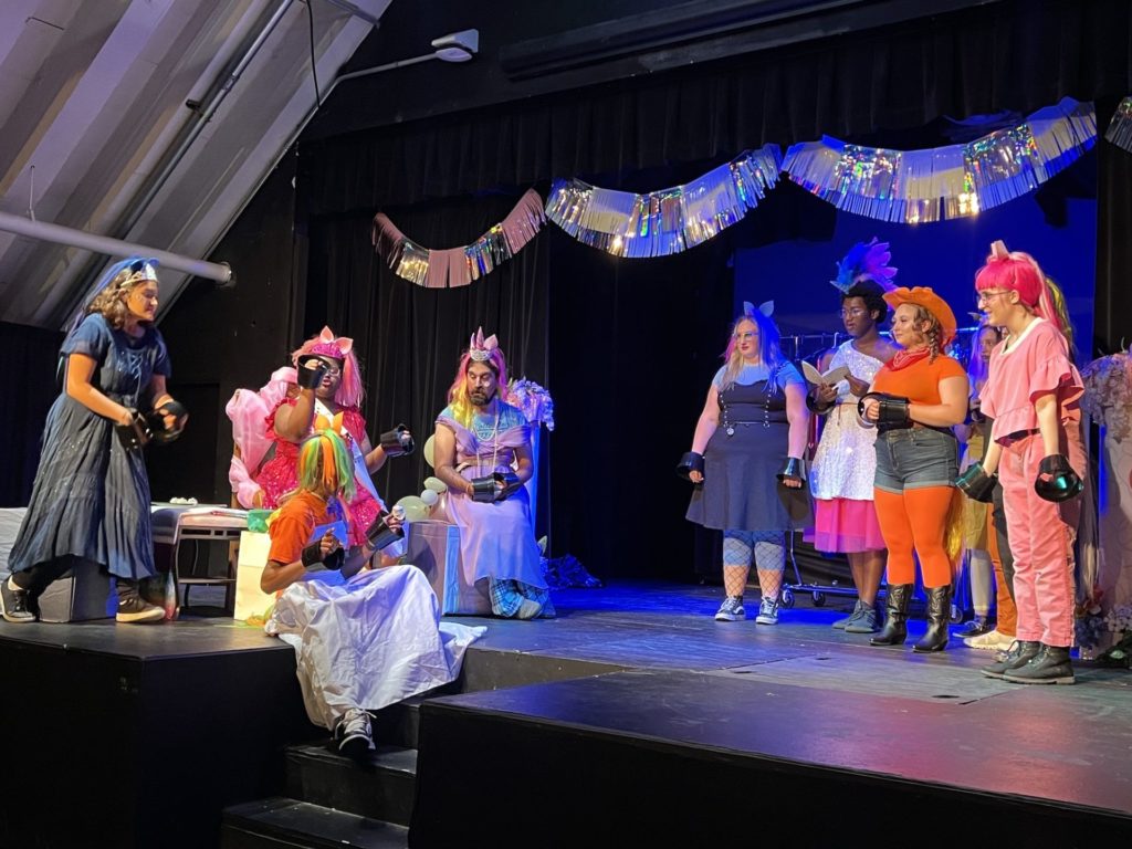 Actors stand on a dimly lit stage that is decorated for a birthday party. On the left is Princess Millenia sitting in a chair with other ponies surrounding her. On the right are the ponies presenting their version of titus andronicus. 