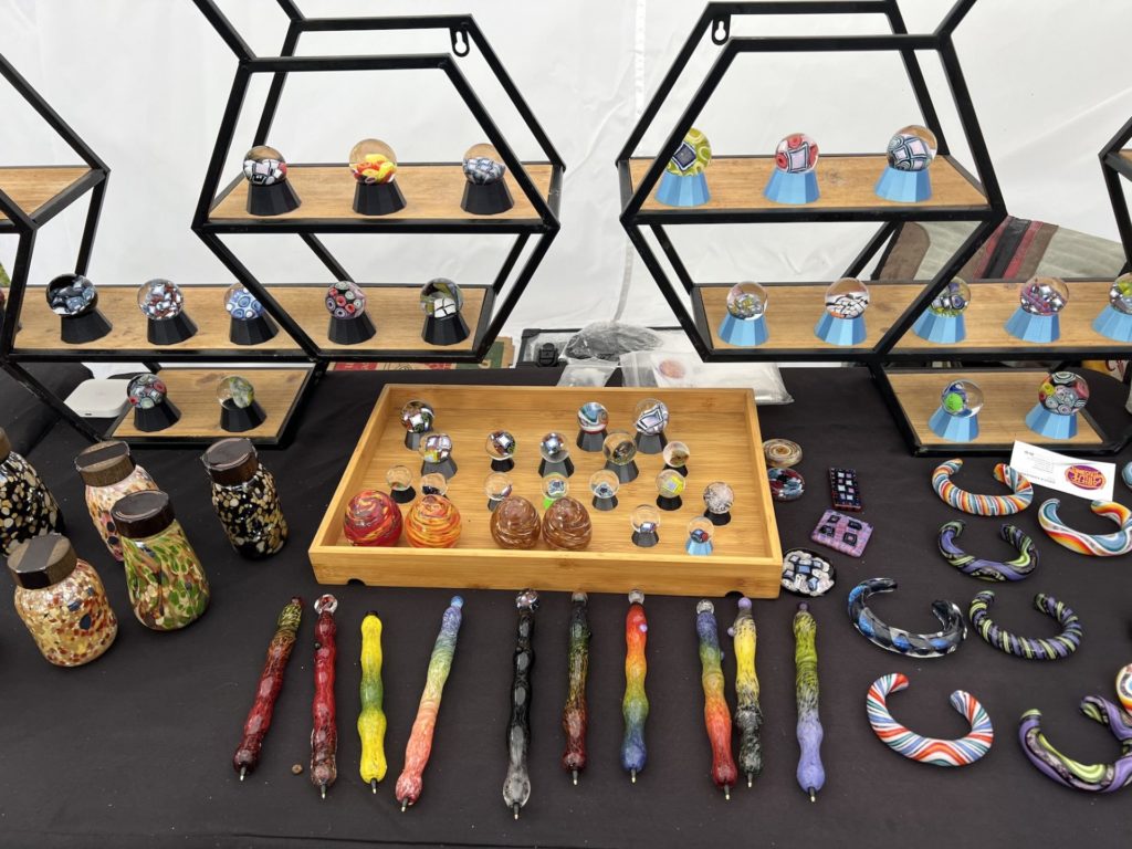 an assortment of glass pieces lay on a table and on hexagon bookshelves; they include small figures, bracelets, jars, mini globes, and blown glass handled pens