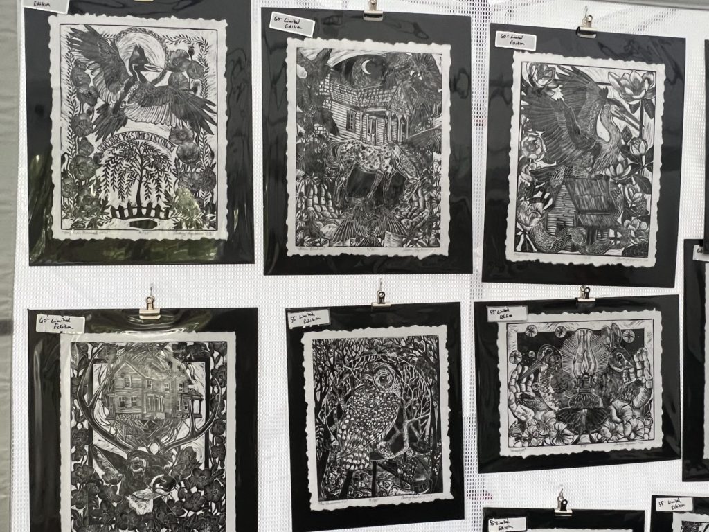 black and white prints hang on a wall in an outdoor tent; they have significant detail and depict various scenes ; mostly combinations of animals and houses (such as a bird sitting on top of a house but the bird is larger than the house)