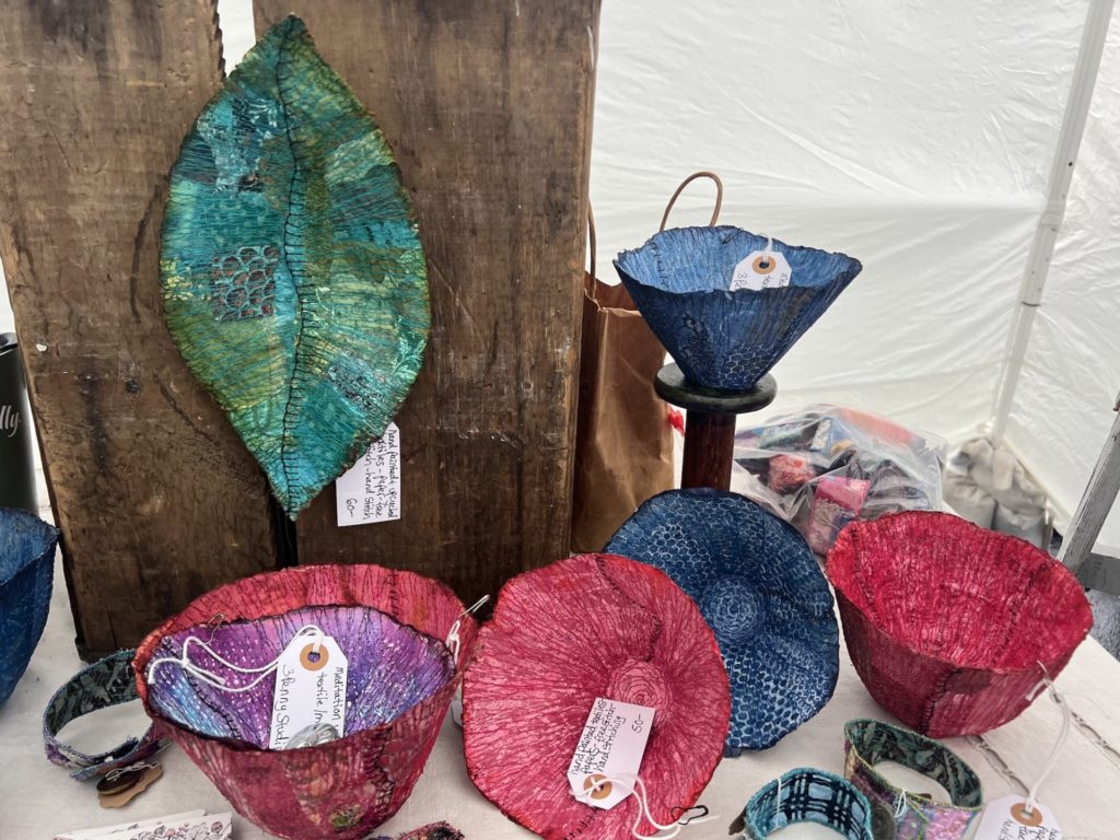 a selection of fiber artwork in bright colors; mostly bowls with a large fiber leaf hanging