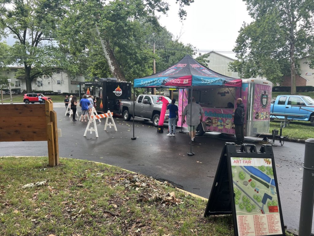 Two food trucks are set up at Crystal Lake Park; the one on the left is dark and serves pizza, the one on the right is bright pink and turquoise with cupcakes depicted on the truck