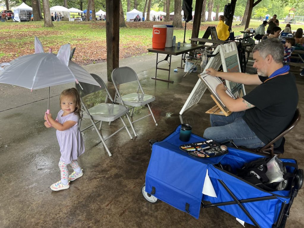 Dan Wild draws an incredibly uncooperative two year old. Wild sits on the right, while a two year girl in a purple dress and leggings runs around holding a unicorn umbrella. 