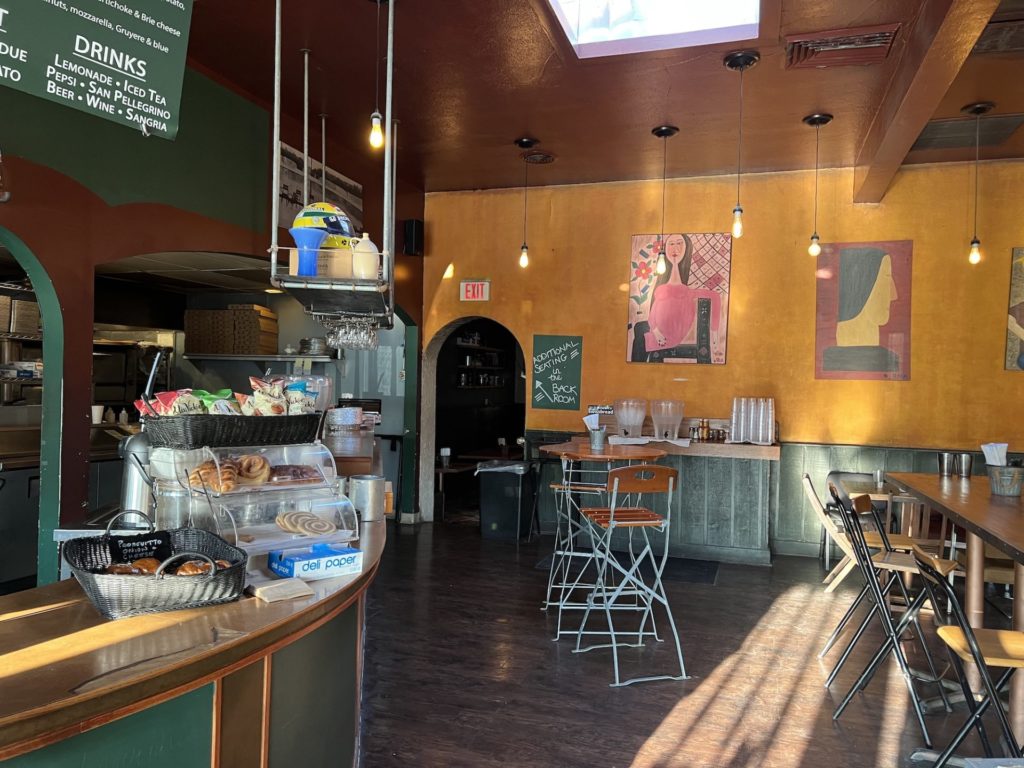 Interior of the bread company is empty. On the left is a counter with a menu hanging above it. The walls are orange with art work. There is an arched doorway leading to more seating. There are tables and a counter with water.