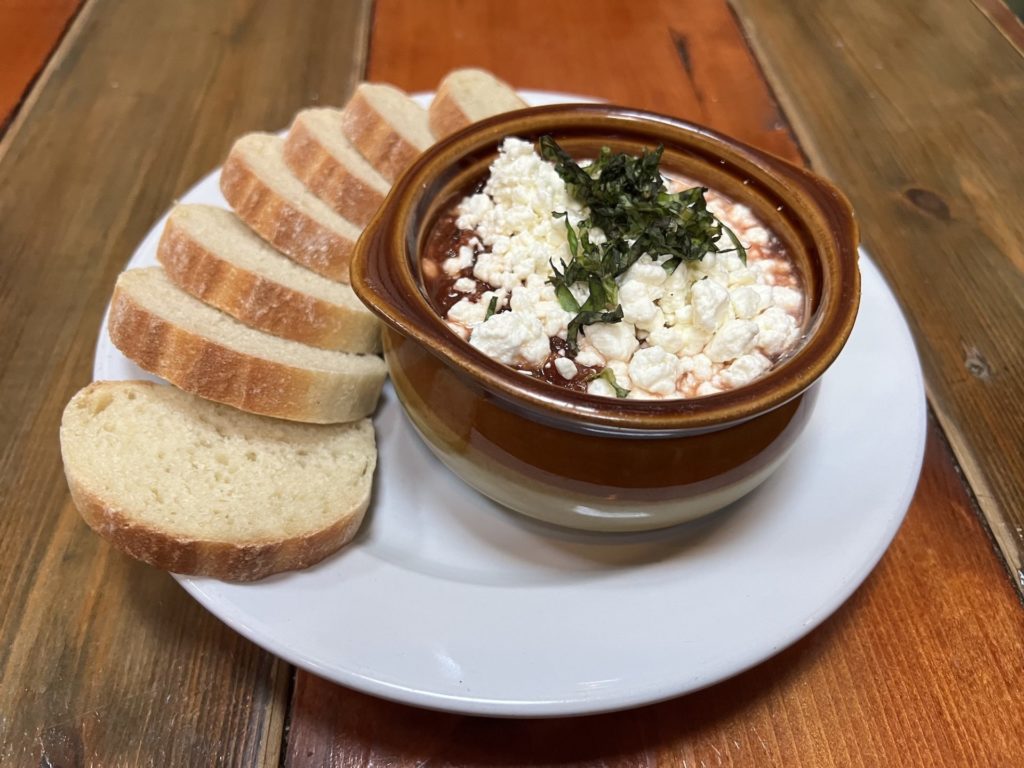 a small round ceramic dish holds a marinara sauce with goat cheese on top. it sits on a white plate on a wooden table. there is a spread of small white slices of bread around the ceramic bowl