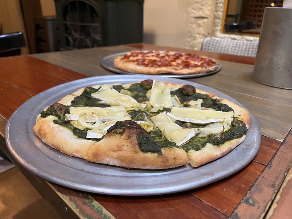 a brie and pesto thin crust pizza on a round metal tray is in the foreground and a matching pepperoni pizza is behind it on a wooden table