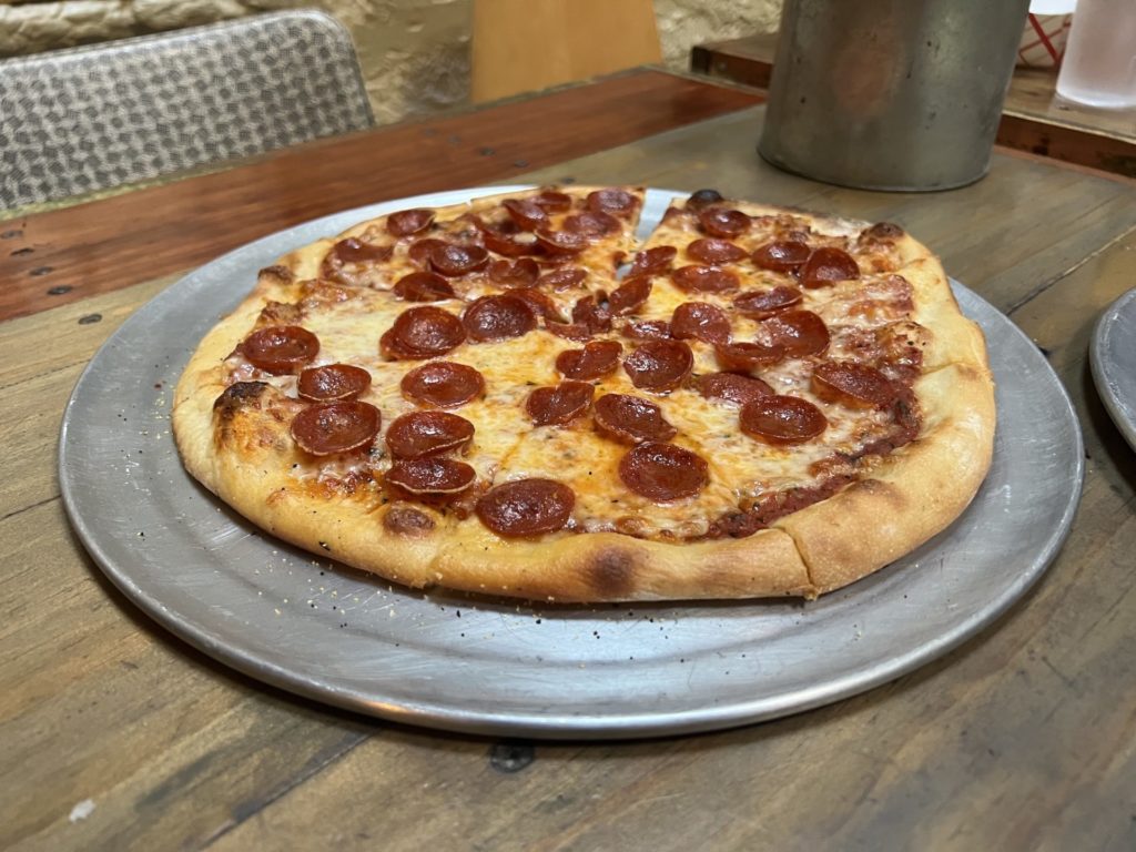 a pepperoni thin crust pizza on a round metal tray sits on a wooden table