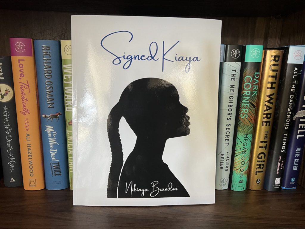 Signed Kiaya stands on a bookshelf leaning against a full shelf of other books. The book is a large softcover. It has a white background with a profile silhouette of a Black woman, with her chin pointed upwards and a long brain hanging from a pony tail.
