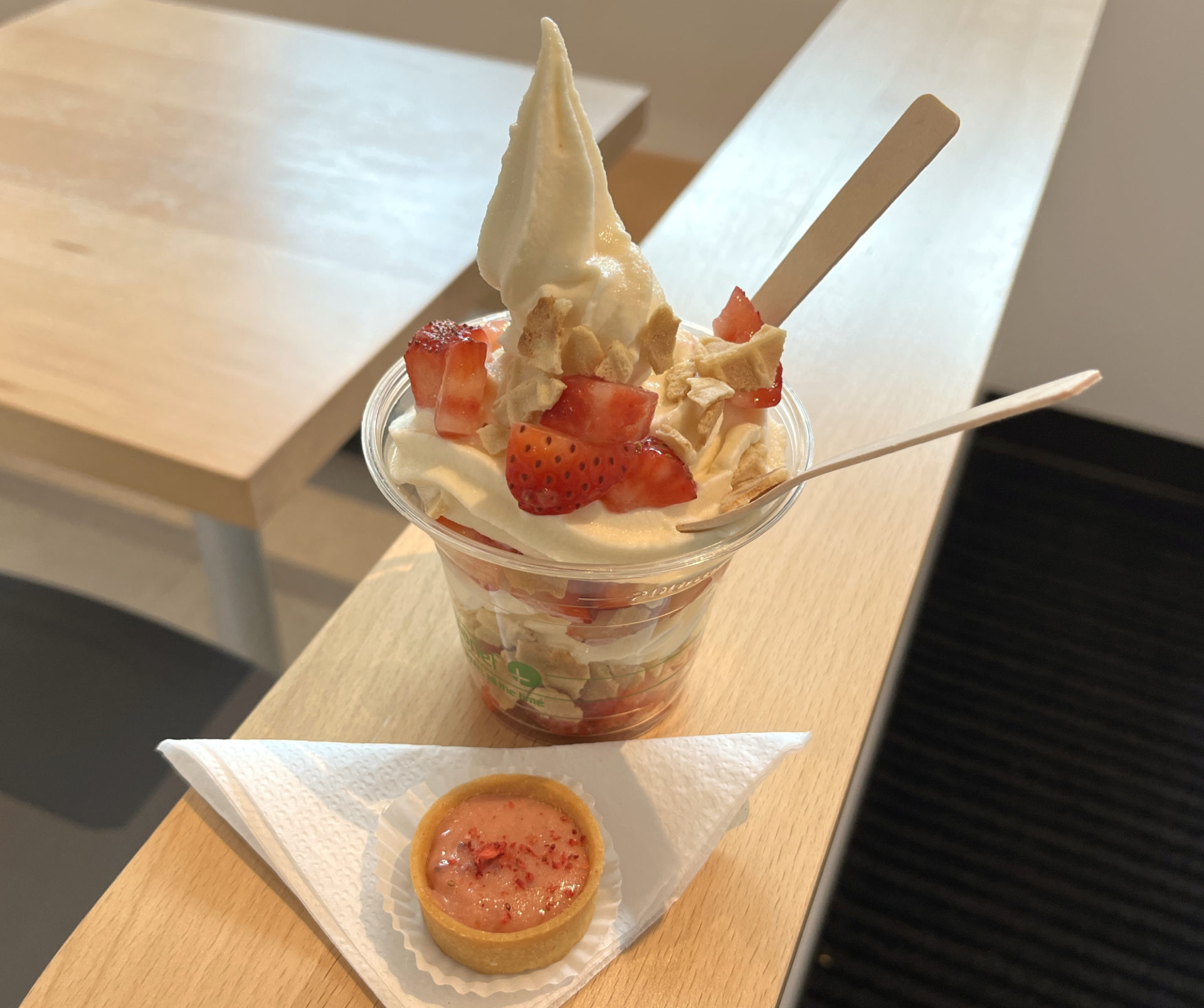A soft serve ice cream parfait in a cup has waffle cone and strawberries on it, with two spoons sticking out. Next to it is a strawberry mini tart, with pink filling, resting on a white paper napkin folded into a triangle. Both items are perched on the light wood top of a ledge.
