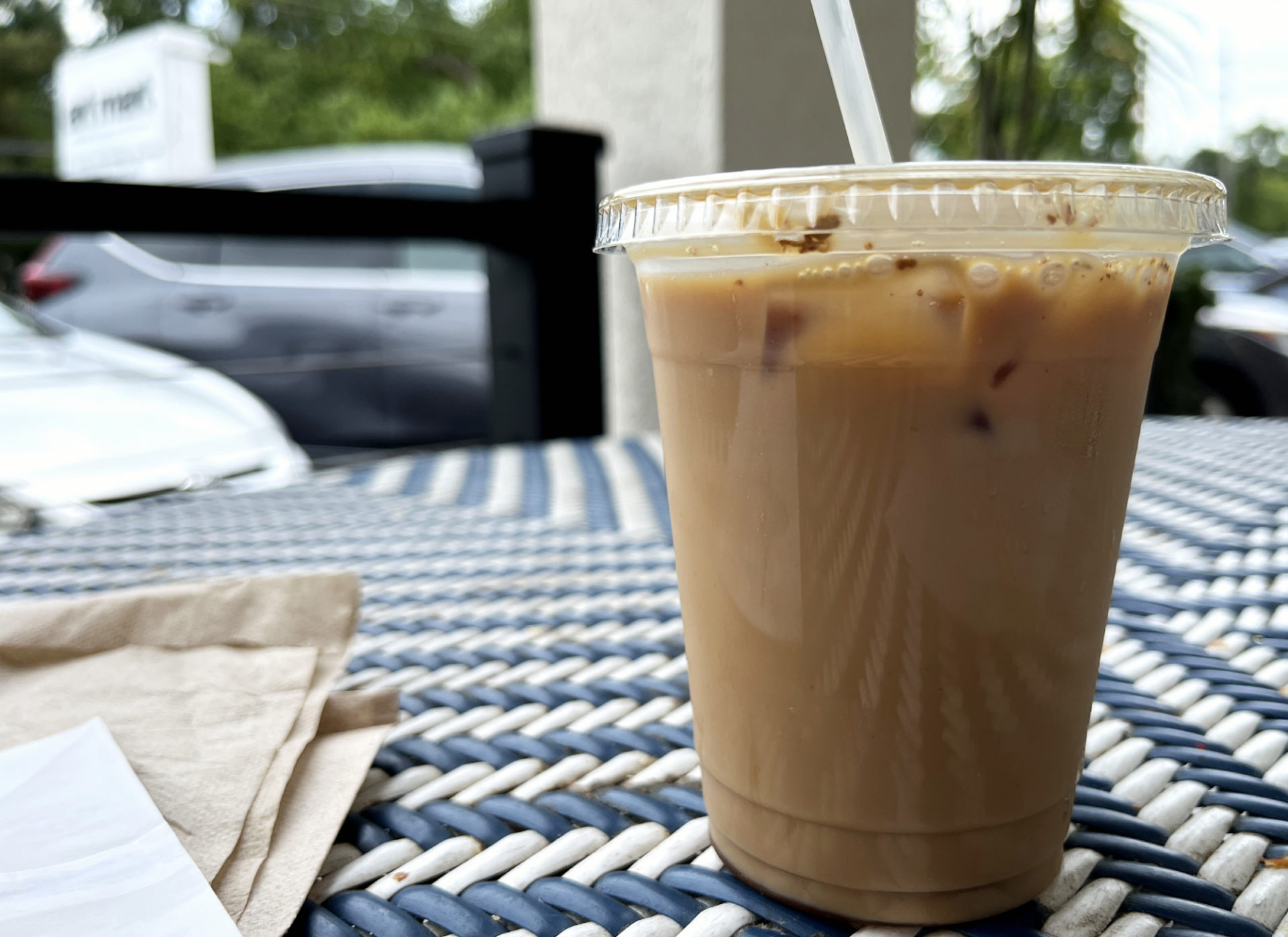 An iced coffee drink in a clear plastic cup is on a blue and white outdoor patio table. To the left of the image are some napkins and papers.
