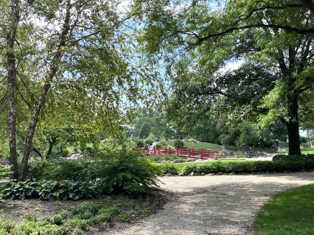 Pathway around the Mabery Gelvin Botanical Gardens, part of the Champaign County Forest Preserve District. There is a red, Japanese-style bridge over a koi point in the middle distance of the photo. Lush trees and a gravel pathway frame the image.