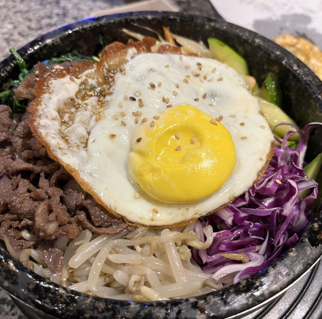 A black stone bowl filled with kimchi bulgogi bibimbap bowl. On top you can see beef, zucchini, red cabbage, sprouts, and a fried egg.