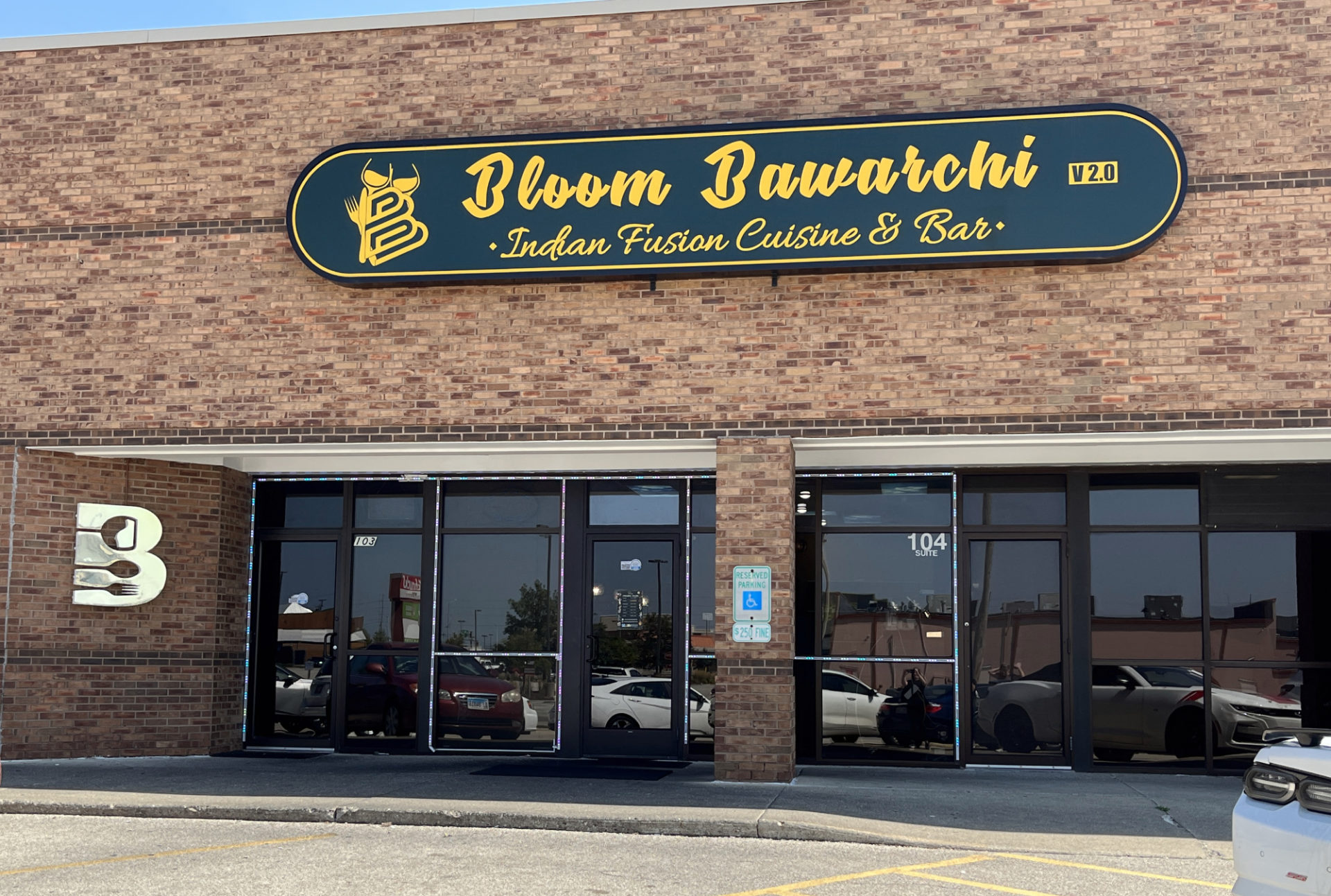 Exterior of Bloom Bawarchi Indian restaurant. A flat, light brown brick facade with a black sign with yellow lettering that reads "bloom bawarchi indian fusion cuisine & bar." Below are eight glass panels, including the entry door.