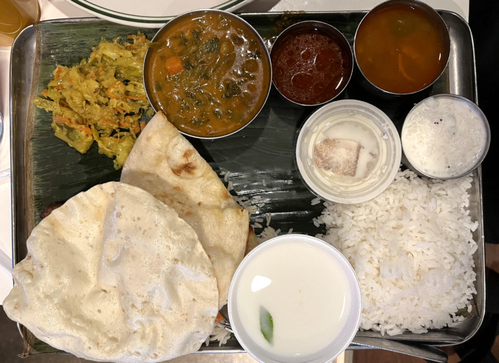 Lunch vegetarian thali at Bloom Bawarchi. A tray covered in a green banana leaf on which four metal bowls of different food are served. Also on the tray is a pile of curried cabbage, naan and papadum, white rice, buttermilk drink, and a milky dessert in a plastic cup.