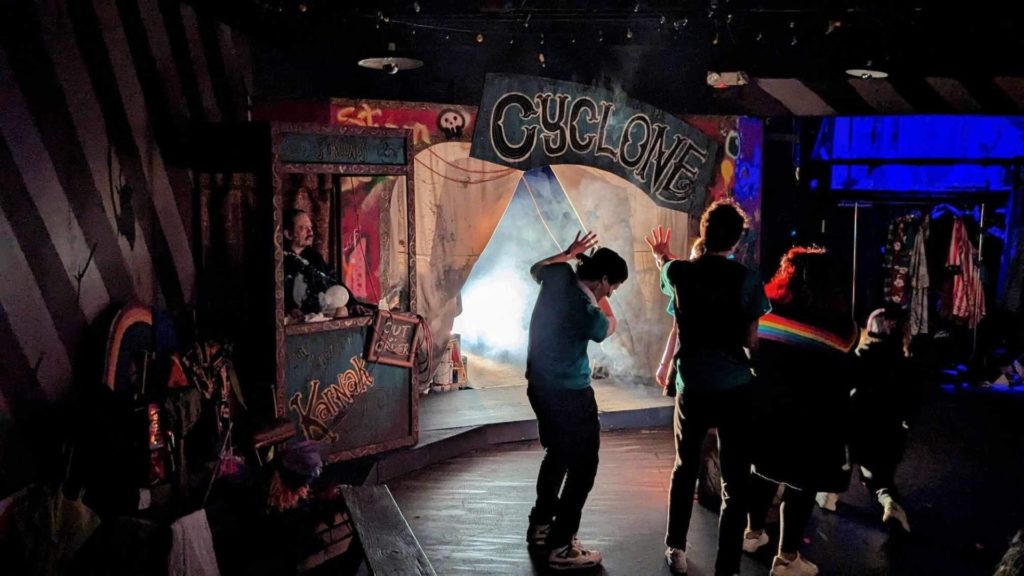 A dark photo of the set of Cyclone the Musical. There is a large sign saying "Cyclone" with a fortune tellers box on the left. there are people standing in the foreground looking at the stage with theirs hands up shielding their eyes from the bright lights from the stage. 