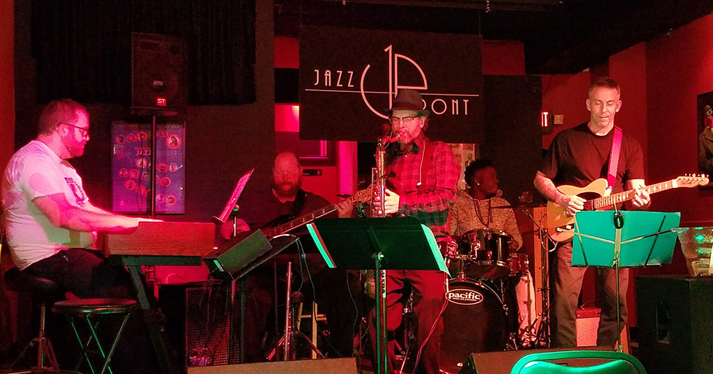 The image is a photo of a jazz band performing on a stage in a dimly lit bar. The stage is decorated with a red curtain and a black banner with “Jazz Up Front” written on it in white letters. The band members are playing various instruments such as a keyboard, drums, saxophone, and guitar, and there are music stands with sheet music in front of them. The foreground consists of empty bar stools and tables. 