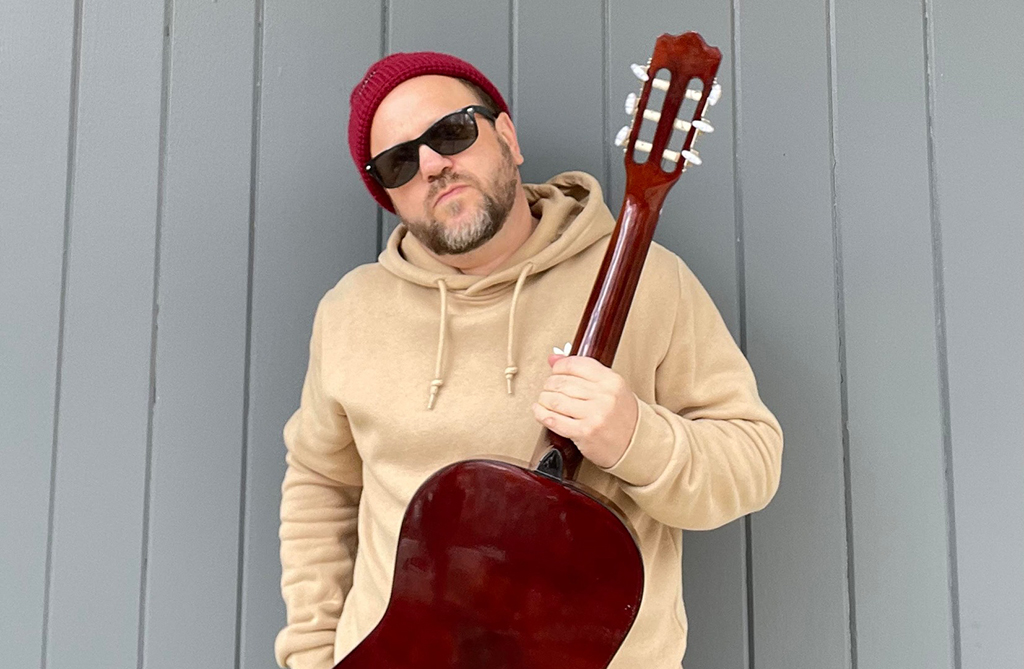 The image you sent is a photo of a person holding a red acoustic guitar. The person is wearing a beige hoodie and a red beanie, and is standing in front of a gray wall with vertical siding