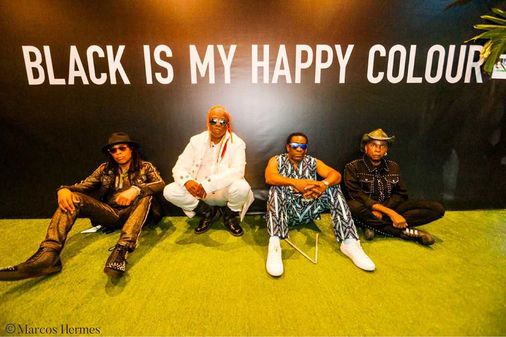 Four people sitting on a green lawn in front of a black wall with the text “BLACK IS MY HAPPY COLOUR” written in white. The person on the left is wearing a black leather jacket and pants, and black boots. The person in the middle left is wearing a white suit and white shoes. The person in the middle right is wearing a blue dress and white shoes. The person on the right is wearing a black and white patterned shirt and black pants.