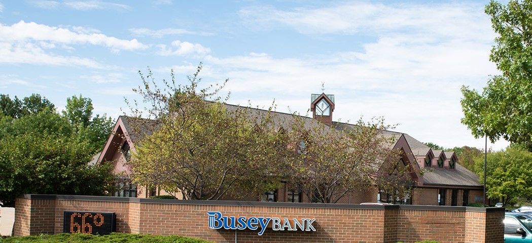 A brick building sits behind a row of small leafy trees, and a low brick wall with a screen indicating the temperature, and Busey Bank in blue and white block letters.