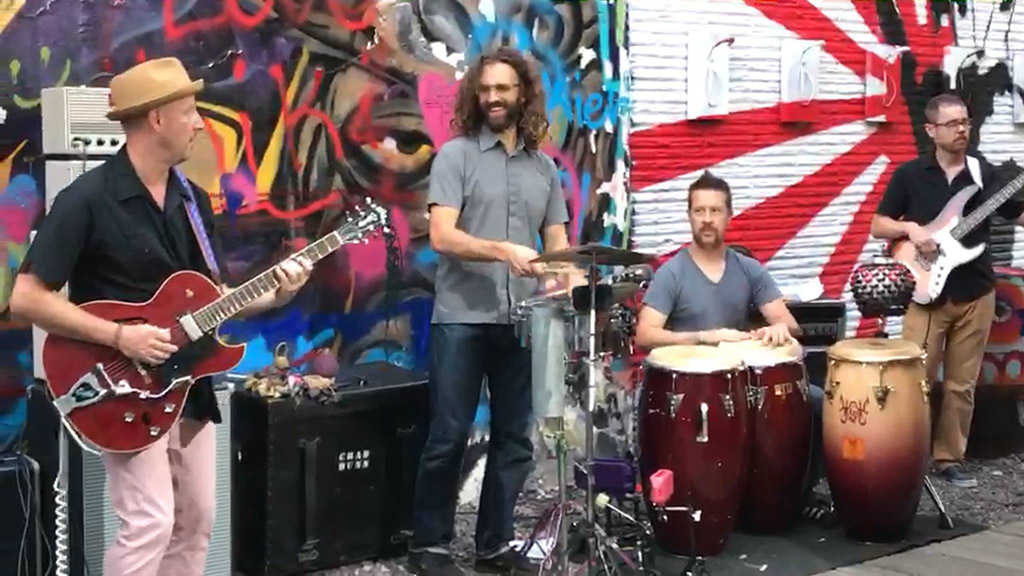 This is a photo of a band playing in front of a colorful graffiti wall. The band consists of a guitarist, a drummer, a percussionist, and a bassist. The guitarist is playing a red electric guitar, the drummer is playing a drum kit, the percussionist is playing congas, and the bassist is playing a white bass guitar. The background consists of a colorful graffiti wall with various designs and patterns.