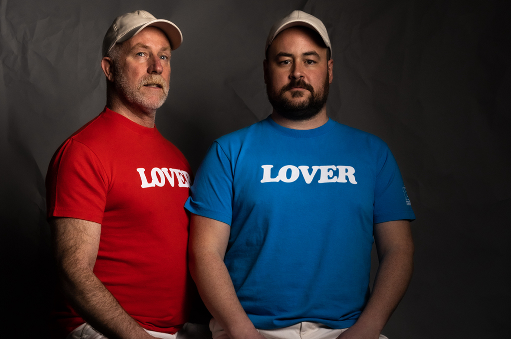This is a photo of two people standing side by side in front of a black background. The person on the left is wearing a red t-shirt with the word “LOVER” written in white letters, while the person on the right is wearing a blue t-shirt with the same word written in white letters. Both people are also wearing baseball caps.