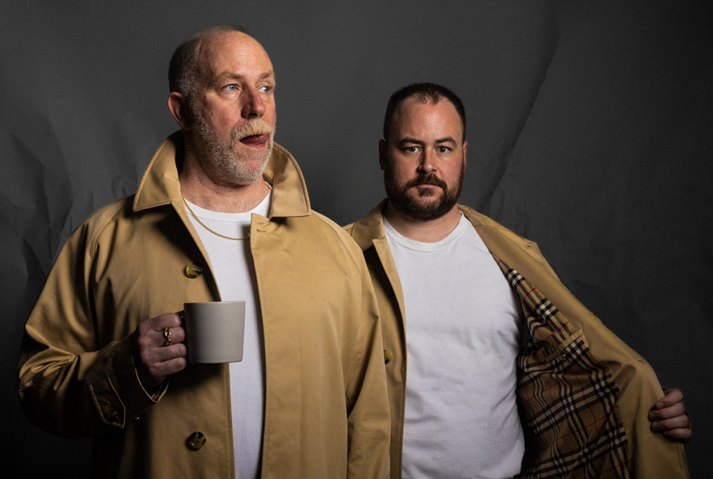 This is a photo-realistic image of two people standing in front of a dark gray background. The person on the left is wearing a yellow raincoat and holding a white mug, while the person on the right is wearing a white t-shirt and a plaid jacket. Both people are standing close together.