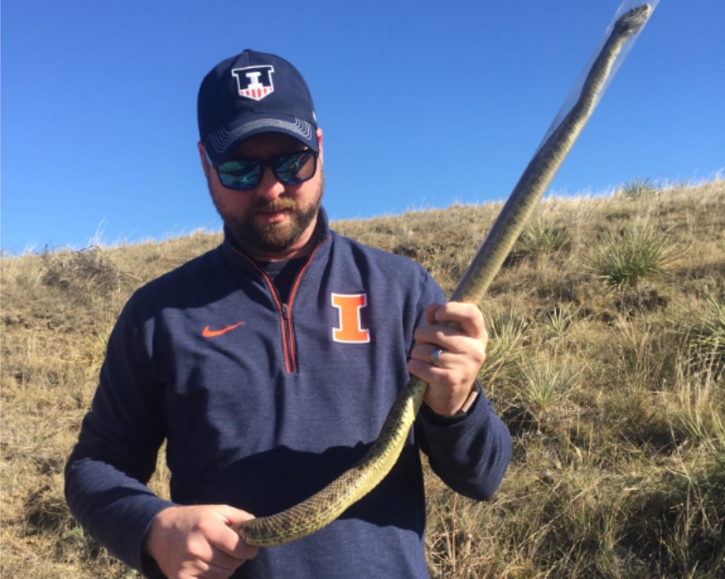 A white man stands in front of a grassy hill and blue sky holding a very long snake. He wears a navy blue hat and shirt with the University of Illinois "I" in orange and white displayed on both. The snake is completely rigid and straight up in the air. 