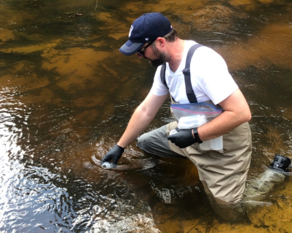 A white man in khaki waders and boots, a white t-shirt, and navy blue hat collects water samples in a body of water. It is brown with big rocks visible at the bottom.