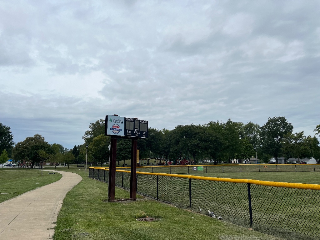A shot of the scoreboard of the McFarland baseball field. There is a winding sidewalk on the left side of the image and the outfield and scoreboard on the right. It's a cloudy day. 