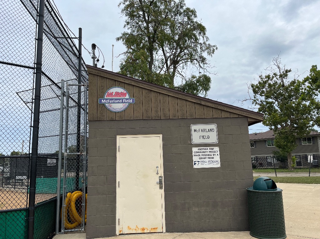 The side view of the McFarland Field concession stand. It is a small gray building with a white door against the tall black baseball fence. 