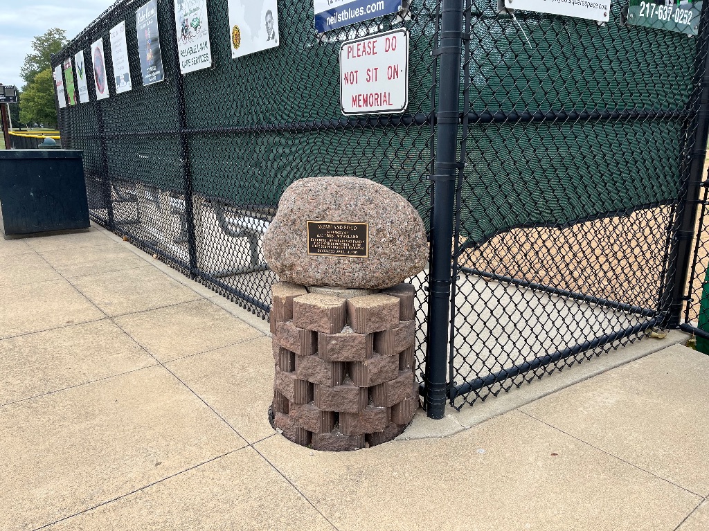 The memorial for Shannon McFarland's grandfather is a large stone with a plaque on top of a circular brick structure. It is sitting in front of a dugout. 