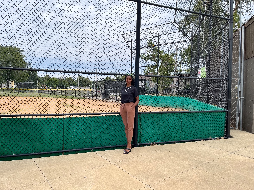 A black woman with long black braids, a black shirt, and tan pants stands in front of a tall black fence with a baseball diamond behind it. There is green padding around the lower part of the fence.