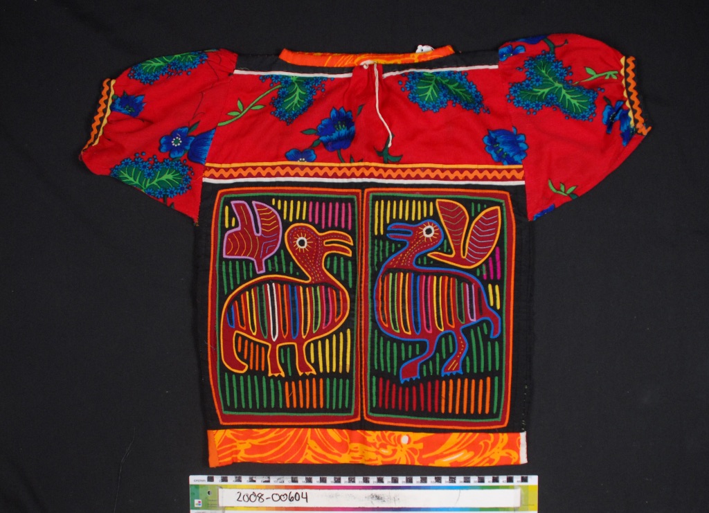 A traditional Mola lays flat against a black background. It is a hand sewn top with giant red flowers, green leaves on the top part and two birds facing each other on the lower part. 