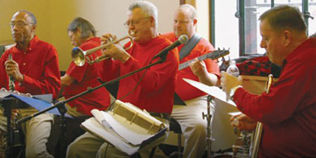 This is a photo of a group of musicians playing various instruments in a room. The musicians are wearing red shirts and are seated on chairs. The instruments being played include a trumpet, a drum set, a guitar, and a keyboard. There are music stands in front of the musicians with sheet music on them. The background consists of a window and a wall with a painting hanging on it.